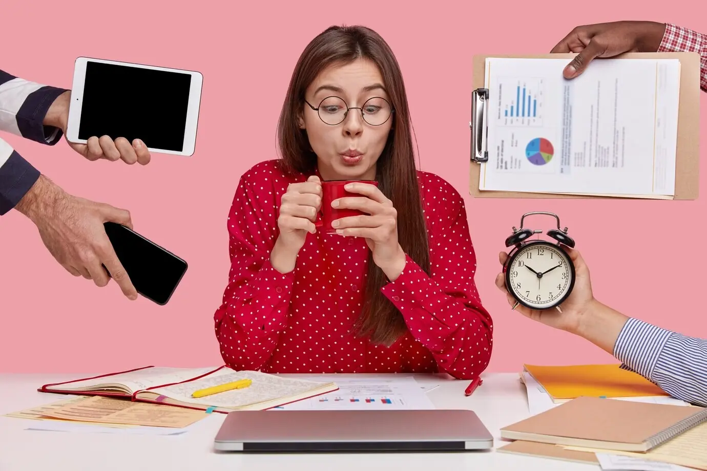 A brunette woman sits at a desk, surrounded by gadgets and papers.