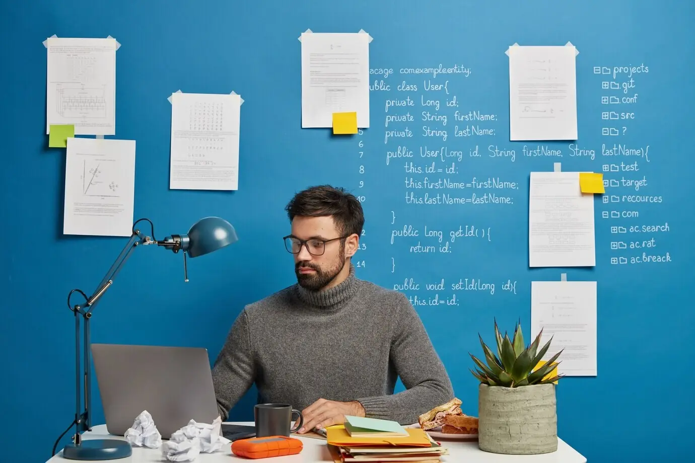 A serious, professional male geek concentrated on the screen of a modern laptop, wearing eyeglasses, posing in a coworking space against a blue background.