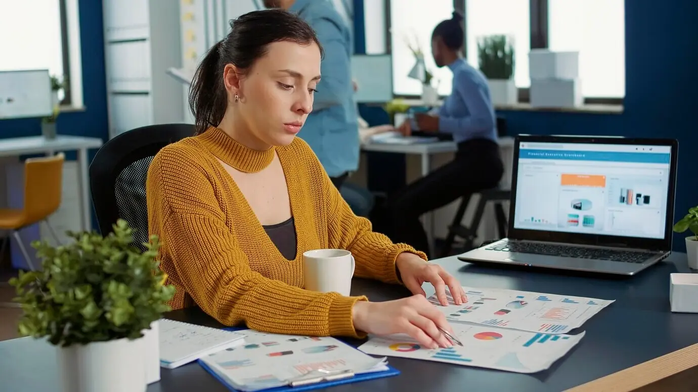 Portrait of a startup employee comparing two sheets of paper with charts to analyze financial data while drinking morning coffee. A woman working in a busy marketing department sits at a desk with a laptop.